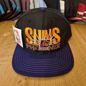 May include: Black and purple snapback hat with the word "SUNS" in gold embroidery. The hat features the Phoenix Suns logo and a purple brim. A tag is attached to the side.