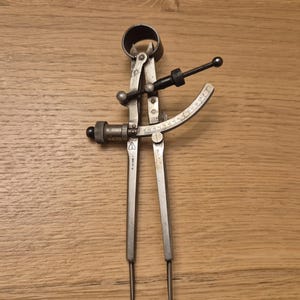 Può includere: Un calibro in metallo vintage con una scala di misurazione curva e bracci regolabili. Lo strumento ha una manopola di regolazione nera e una parte superiore circolare, probabilmente utilizzata per misurazioni precise. Il calibro è in metallo argentato.