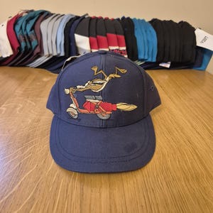 以下が含まれることがあります： ネイビーブルーの野球帽。赤いスクーターに乗ったワイリー・コヨーテの刺繍が施されています。帽子はカーブしたつばと構造化されたクラウンが特徴です。さまざまな野球帽のコレクションが背景に見えます。