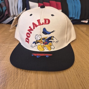 Könnte beinhalten: Weiß-schwarze Baseballkappe mit gesticktem Donald Duck-Motiv. Das Wort "DONALD" ist rot auf der Vorderseite gestickt. Im Hintergrund sind weitere Kappen zu sehen. Die Kappe hat einen flachen Schirm.
