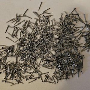 May include: A collection of small, silver-coloured nails scattered on a light surface. The nails have flat heads and pointed tips, suitable for woodworking or construction. The nails vary in length.