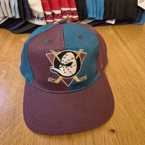 Op de afbeelding: Een bordeauxrode en blauwgroene baseballcap met het logo van een hockeyteam met een eend-ontwerp en gekruiste hockeysticks. Andere caps zijn zichtbaar op de achtergrond.