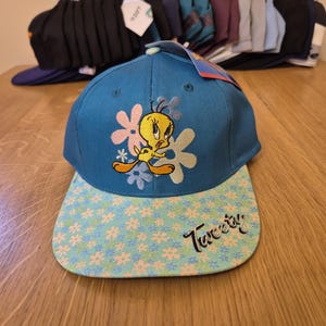 Può includere: Un cappellino da baseball blu con Tweety Bird e dettagli floreali. Il cappellino ha una visiera azzurra con un motivo floreale e la scritta "Tweety" in nero. Altri cappellini sono visibili sullo sfondo.