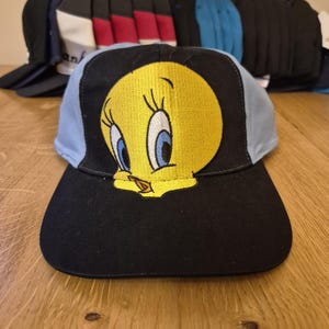Può includere: Un cappellino da baseball nero con visiera azzurra e un ricamo di Titti giallo sul davanti. Altri cappellini da baseball sono visibili sullo sfondo, con vari colori e disegni.