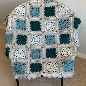 May include: A crocheted blanket with a square pattern in shades of white, blue, and green. The blanket is folded and draped over a black metal chair.