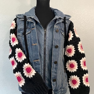 May include: A denim jacket with a zip-up front and a hood. The sleeves are crocheted with a black background and white and pink flower patterns. The jacket is displayed on a black mannequin.