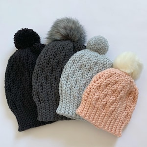 Op de afbeelding: Vier gebreide mutsen in verschillende kleuren en maten. De mutsen zijn allemaal gemaakt met een textuur en hebben pompoenen bovenop. De mutsen zijn gerangschikt in een rij van de grootste tot de kleinste, waarbij de grootste muts zwart is, de op een na grootste grijs, de volgende lichtblauw en de kleinste roze.