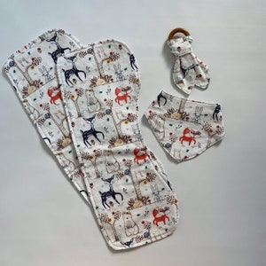 May include: A set of three baby bibs and a teether with a woodland animal print. The bibs are white with a brown, red, and blue pattern of deer, foxes, bears, and rabbits. The teether is made of wood and has a brown color.