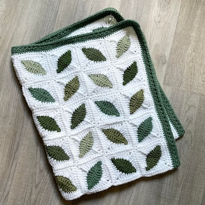 Puede incluir: Una manta de crochet blanca con un patrón de hojas verdes. La manta está hecha con un punto sencillo y tiene un borde de crochet verde.