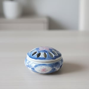 Op de afbeelding: Een kleine keramische wierookbrander met een blauw-wit bloemmotief. Het deksel heeft verschillende ovale openingen. De brander is versierd met roze en blauwe bloempatronen. De achtergrond is wazig.