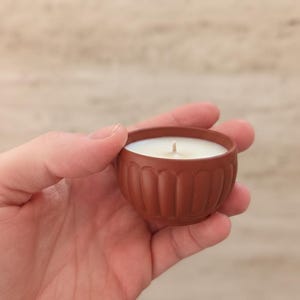 Peut inclure: Une petite bougie de couleur terre cuite dans un bol décoratif. La bougie a une cire blanche et une seule mèche. Le bol a un design côtelé à l'extérieur. La bougie est tenue dans une main.