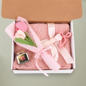 May include: A gift box containing a pink cable-knit blanket, a small glass candle with a floral label, and a bouquet of three crocheted tulips in pink and light pink with green stems, all tied together with a light pink satin ribbon.