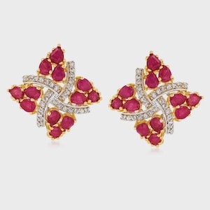May include: A pair of gold-toned stud earrings. Each earring features a floral design with ruby-colored gemstones and small, clear gemstones. The earrings are set against a light background.