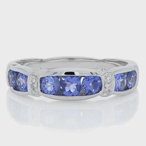 May include: A silver ring featuring five oval, blue gemstones. Small, clear stones are set between the blue gems. The ring is displayed on a white background, reflecting the light.