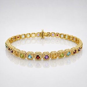 May include: A gold-toned bracelet featuring a series of square-cut gemstones in various colors, including red, blue, green, and purple. The bracelet has an intricate filigree design around each stone and a secure clasp.