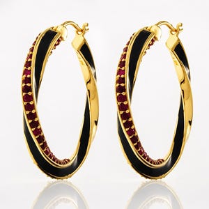 May include: A pair of gold-colored hoop earrings. Each earring features a twisted design with a black enamel stripe and a row of small, red gemstones. The earrings are displayed against a white background.