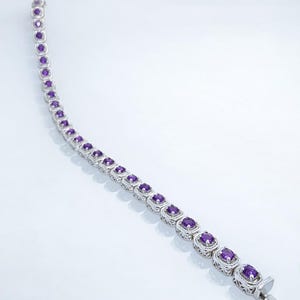 Pode incluir: Uma pulseira de prata com pedras preciosas roxas de corte quadrado, engastadas em molduras ornamentadas e vazadas. A pulseira tem um fecho e é exibida sobre um fundo branco. A joia tem aproximadamente 18 cm de comprimento.