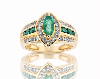 May include: A gold ring featuring a marquise-cut emerald center stone, surrounded by a halo of diamonds. Additional emeralds and diamonds are set along the band. The ring is displayed on a reflective surface.