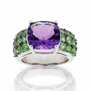 May include: A silver ring featuring a large, square-cut, purple amethyst gemstone as the centerpiece. The ring is flanked by clusters of small, round, green gemstones, creating a vibrant contrast. The ring is set against a white background.