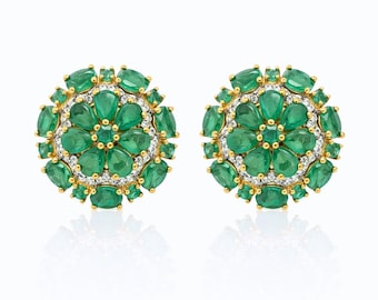 May include: A pair of gold-toned stud earrings. Each earring features a floral design composed of emerald-green gemstones and small white gemstones. The earrings are set against a white background.