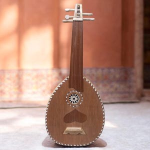 Op de afbeelding: Een houten oed, een snaarinstrument uit het Midden-Oosten, wordt getoond. De peervormige body heeft een decoratieve rozet en hartvormige klankgaten. Het instrument heeft een lange hals met stemschroeven en is afgewerkt met witte accenten.