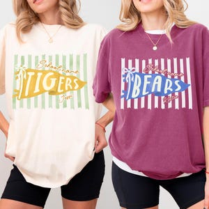 May include: Two short-sleeve crewneck t-shirts, one ivory and one berry, each featuring a central graphic of a vertical striped background with a pennant banner. The ivory shirt displays the text TIGERS, and the berry shirt displays the text BEARS.