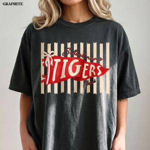 May include: A graphite-colored short-sleeve t-shirt featuring a graphic of a red pennant flag with the text "TIGERS" in white. The pennant is accented with a white bow and sits over a background of vertical beige stripes. The word "GRAPHITE" is printed in the top left corner.