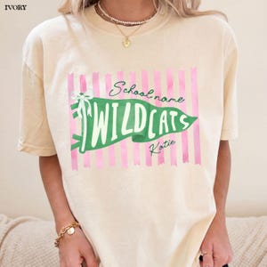 May include: An ivory short-sleeve t-shirt featuring a graphic of a green pennant flag with a white bow. The flag displays the text "School name", "WILDCATS", and "Katie" over a background of vertical pink stripes. The shirt has a crew neckline.