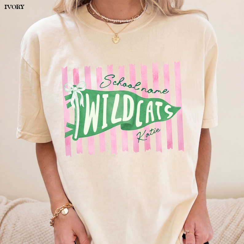 May include: An ivory short-sleeve t-shirt featuring a graphic of a green pennant flag with a white bow. The flag displays the text "School name", "WILDCATS", and "Katie" over a background of vertical pink stripes. The shirt has a crew neckline.