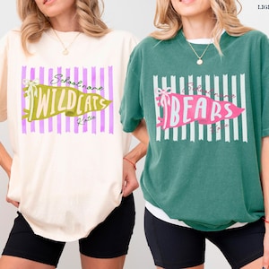 May include: Two short-sleeve crewneck t-shirts, one ivory and one light green. The ivory shirt features a purple striped background with a green pennant graphic reading Wildcats. The green shirt has white stripes and a pink pennant reading Bears.
