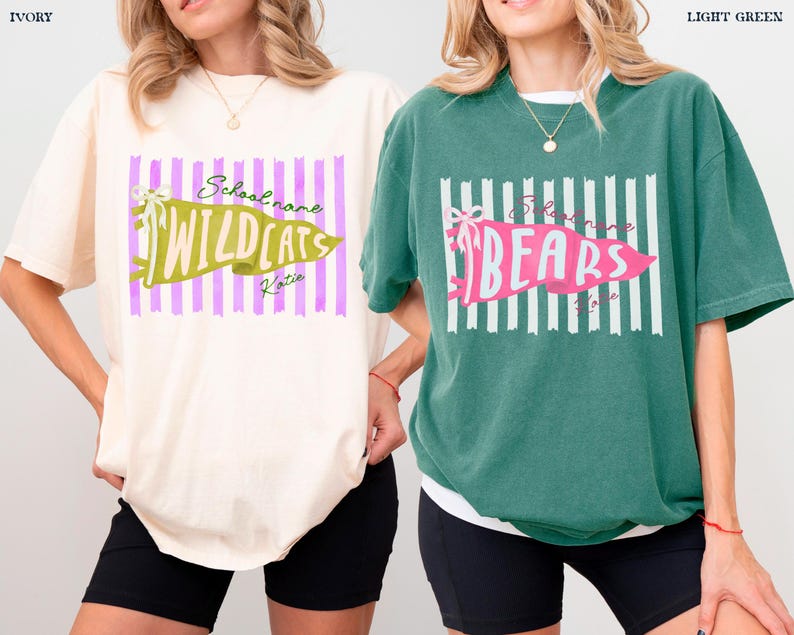 May include: Two short-sleeve crewneck t-shirts, one ivory and one light green. The ivory shirt features a purple striped background with a green pennant graphic reading Wildcats. The green shirt has white stripes and a pink pennant reading Bears.