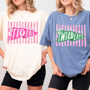 May include: An ivory t-shirt and a washed denim blue t-shirt, both featuring a graphic of pink vertical stripes, a pennant with a bow, and text reading School name, TIGERS, Jessi, and School name, WILDCATS, Katie, with short sleeves and crew neck.