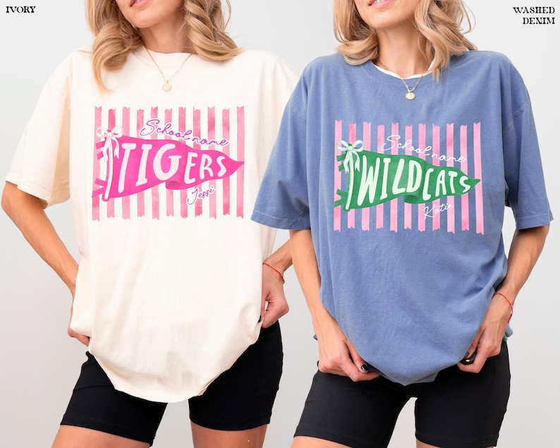 May include: An ivory t-shirt and a washed denim blue t-shirt, both featuring a graphic of pink vertical stripes, a pennant with a bow, and text reading School name, TIGERS, Jessi, and School name, WILDCATS, Katie, with short sleeves and crew neck.