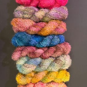 May include: A selection of vibrant yarn skeins in a range of colours, including pink, purple, blue, and yellow. The yarn has a textured, fluffy appearance, suitable for knitting or crochet. The skeins are neatly stacked against a dark backdrop.