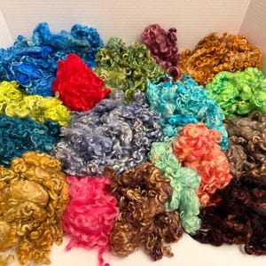 Op de afbeelding: Een verzameling kleurrijke, krullende wolroving in verschillende tinten, waaronder blauw, groen, rood en bruin. De wol is in een doos gerangschikt en toont de textuur en levendige kleuren van de vezel. Ideaal voor vezelkunstprojecten.