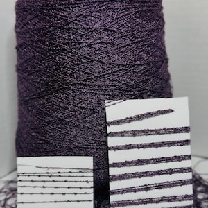 May include: A large spool of purple plum yarn with a textured surface. Two white cards display samples of the yarn, showing horizontal lines. The text "Purple Plum" is visible. A measuring tape is at the bottom.
