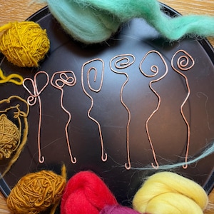 Puede incluir: Una bandeja negra contiene horquillas de alambre de cobre con diseños en espiral y bucle. Ovillos de hilo amarillo y marrón, junto con mechones de lana verde, roja y morada, sugieren un tema artesanal.