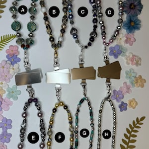 May include: A collection of eight beaded phone charms in various designs. The charms feature a variety of beads in colors like teal, purple, gold, and silver. Each charm has a metal clip for attaching to a phone.