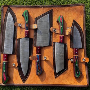 Puede incluir: Un juego de seis cuchillos de cocina con mangos de madera de colores, en un estuche de cuero. Las hojas son plateadas y los mangos tienen tonos verdes, rojos y azules. El estuche es de color marrón claro con costuras marrones y correas de cuero.