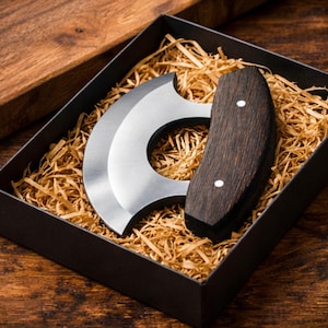 Könnte beinhalten: Ein Mezzaluna-Messer aus Edelstahl mit dunklem Holzgriff, präsentiert in einer schwarzen Geschenkbox mit Stroh. Die gebogene Klinge ist zum Hacken von Kräutern und Gemüse konzipiert.