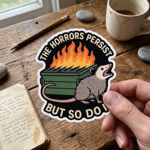 May include: A sticker with the text "THE HORRORS PERSIST BUT SO DO I" features a cartoon opossum next to a dumpster on fire. The sticker is held by a hand, with a journal and coffee cup in the background.