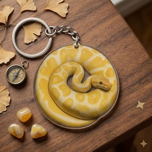 May include: A round keychain featuring a coiled yellow and white snake design. The keychain has a silver chain and ring. Additional items include a small compass, yellow stones, and dried leaves.