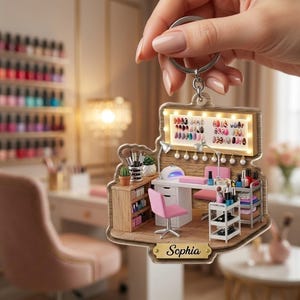 May include: A decorative acrylic keychain featuring a miniature 3D-style nail salon scene with a desk, pink chair, nail polish rack, and a lighted display board. The base includes a gold-toned nameplate with the name Sophia printed in black.