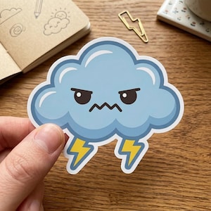 May include: A sticker featuring a cartoon cloud with an angry expression. The cloud is light blue with a white outline and two yellow lightning bolts. The sticker is held by a hand.