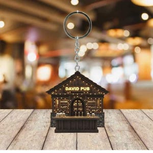 May include: A miniature keychain shaped like a pub, with the text "DAVID PUB" above the entrance. The wooden structure features a bar with beer taps and mugs. The keychain is suspended from a silver ring and chain, with a blurred background.