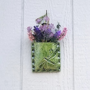 May include: A green ceramic wall planter with a dragonfly design, holding a bouquet of purple and pink flowers.