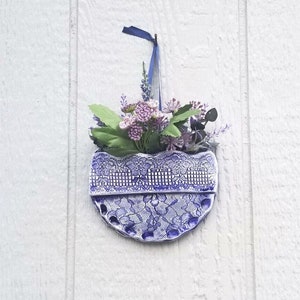 May include: A blue and white ceramic planter with a lace design hangs on a white wall. The planter is filled with purple flowers and green leaves.