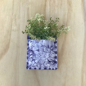 Puede incluir: Un macetero de pared de cerámica morado con un diseño floral. El macetero tiene una forma cuadrada y una pequeña abertura en la parte superior para plantar. Una pequeña planta con flores blancas está creciendo en el macetero.