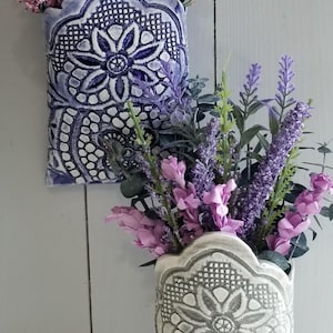 May include: Two decorative wall vases, one blue and one gray, filled with purple and pink flowers. The vases have a detailed floral pattern. The background is a light gray wooden wall.