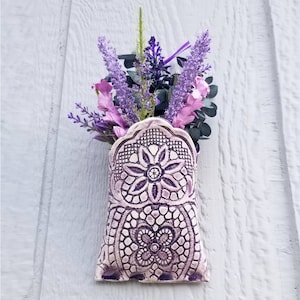 Puede incluir: Un jarrón de pared de cerámica con un diseño de encaje morado y blanco. El jarrón está lleno de flores moradas y rosas.